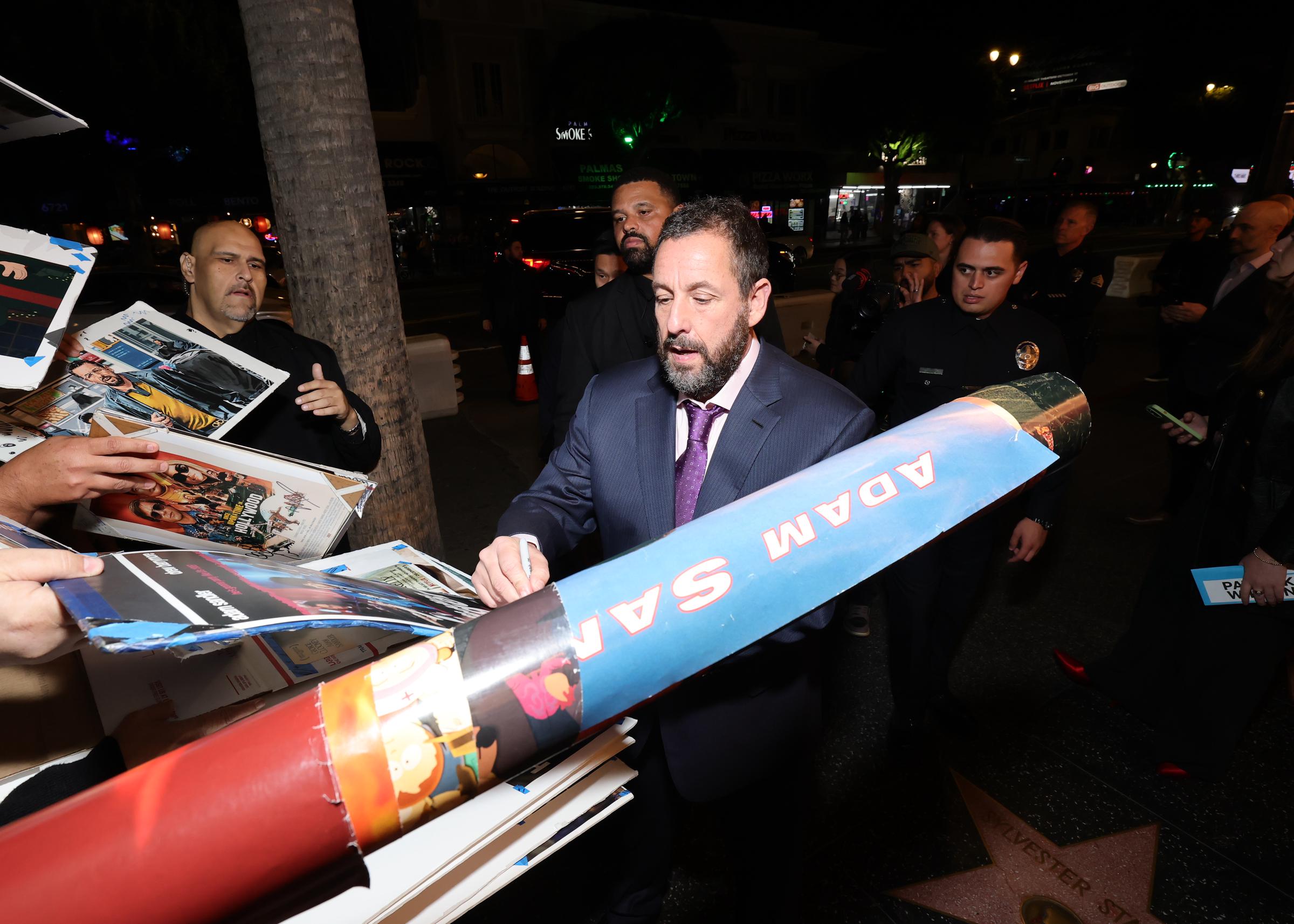 Adam Sandler signs autographs as several fans gather at Netflix’s "Jay Kelly" Los Angeles premiere. His security looks on as the actor warmly attends to the movie lovers.