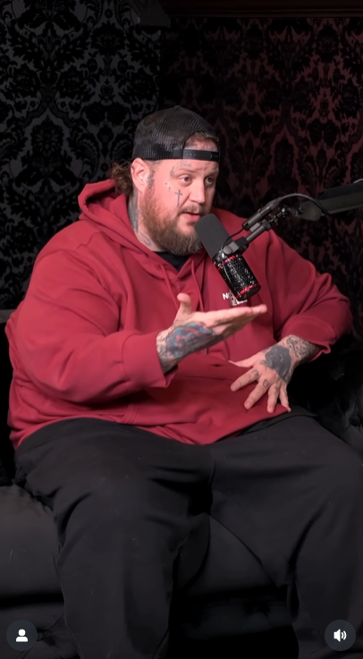 Jelly Roll wears a red hoodie and black cap while talking and gesturing during the "Dumb Blonde" podcast, as posted in October 2025 | Source: Instagram/officialdumbblonde