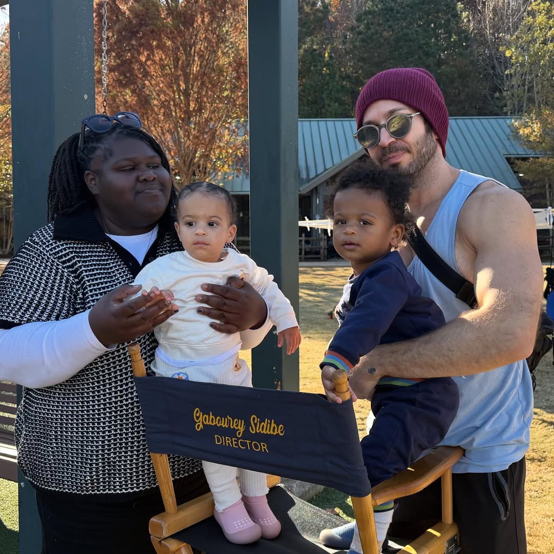 Standing side by side, Gabourey Sidibe and Brandon Frankel face forward, each holding one twin as they rest their hands on Sidibe's director’s chair, centered between them. | Source: Instagram/brandontour