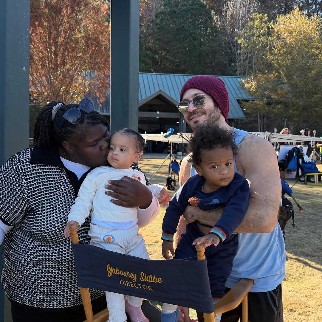 Gabourey Sidibe kisses Maya's cheek while holding her upright; Brandon Frankel stands next to them, angled toward the camera, gently supporting Cooper in one arm. | Source: Instagram/brandontour
