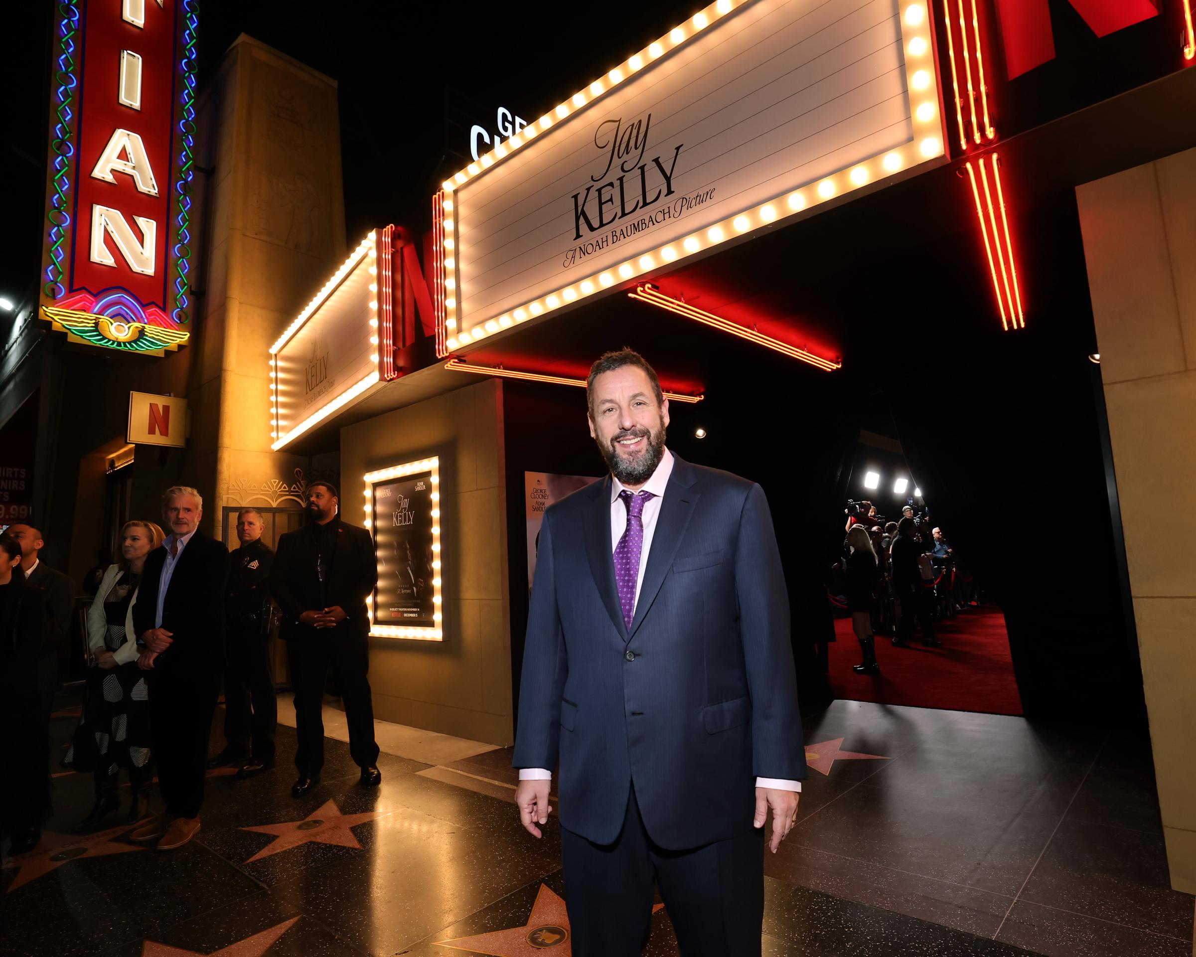 Adam Sandler wears a happy, proud smile as he attends Netflix’s "Jay Kelly" premiere on November 11, 2025, in Los Angeles, California. He poses in front of the brightly lit Egyptian Theatre, exuding confidence.