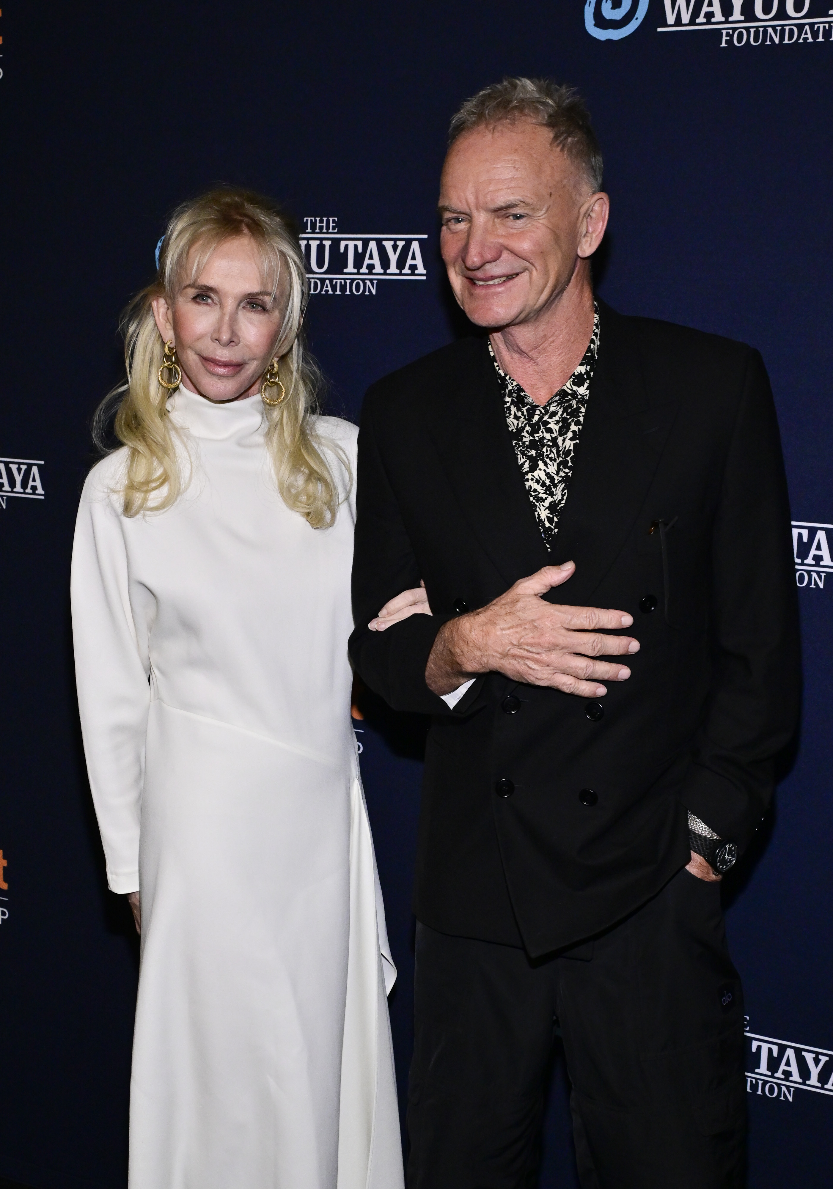 Trudie Styler wears a high-neck, long-sleeved white gown and large dangling golden hoop earrings, while attending The Wayuu Taya Foundation's Spring Gala with Sting at Chelsea Industrial on March 11, 2025, in New York City.